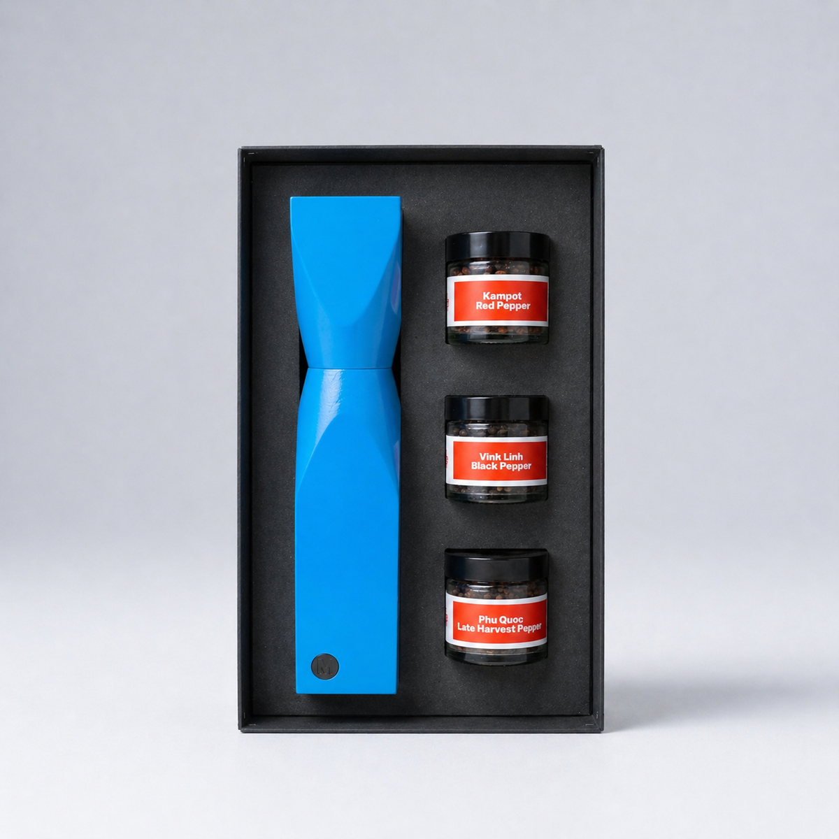 Set of three small jars with red labels and a blue peppermill in a black box on a light gray background