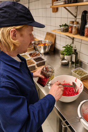 A person in a navy blue work jacket and cap stands at a kitchen counter, holding a glass jar filled with red food while mixing shredded red ingredients in a white bowl. Shelves with jars and kitchen items are visible in the background.