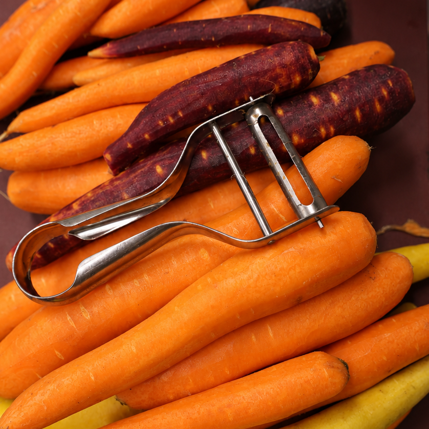 A pile of assorted vegetables on a dark cutting board, including orange carrots, purple sweet potatoes, yellow turnips, and a metal peeler.